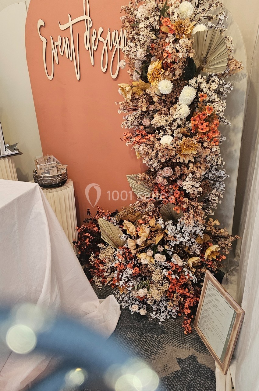 Mur décoratif avec fleurs séchées et fraîches dans des tons chauds, accompagné d'un panneau ’Event design’.