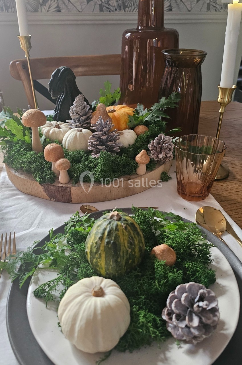 Centre de table automnal avec citrouilles, pommes de pin, mousse et champignons décoratifs sur une table dressée.