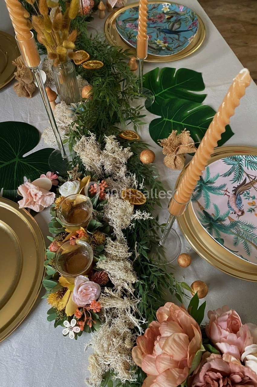 Table décorée avec des assiettes dorées, des fleurs, des bougies torsadées et un chemin de table végétal avec des touches…