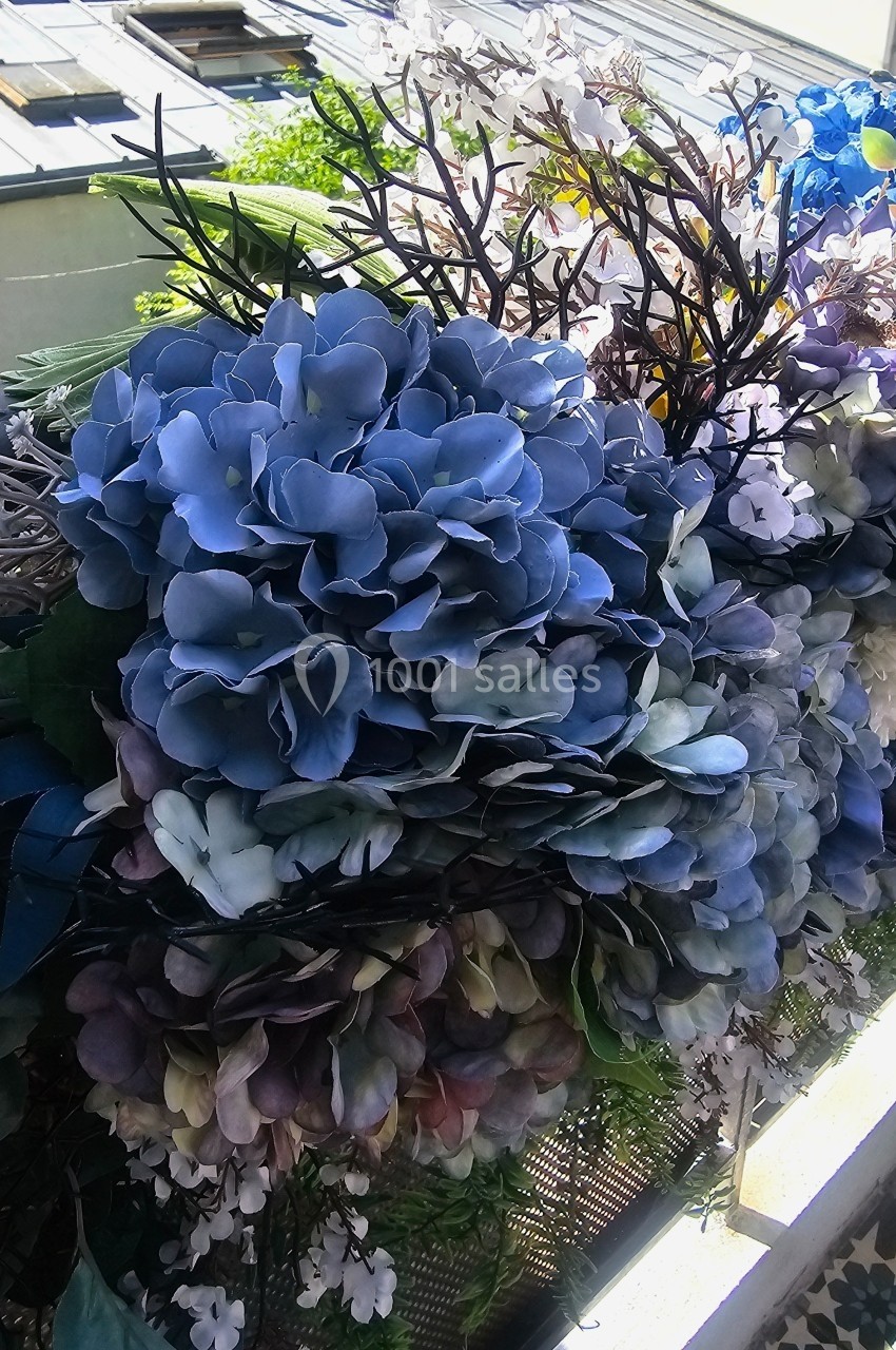 Fleurs artificielles aux tons bleus et violets disposées sur un balcon avec vue sur des toits et de la végétation.