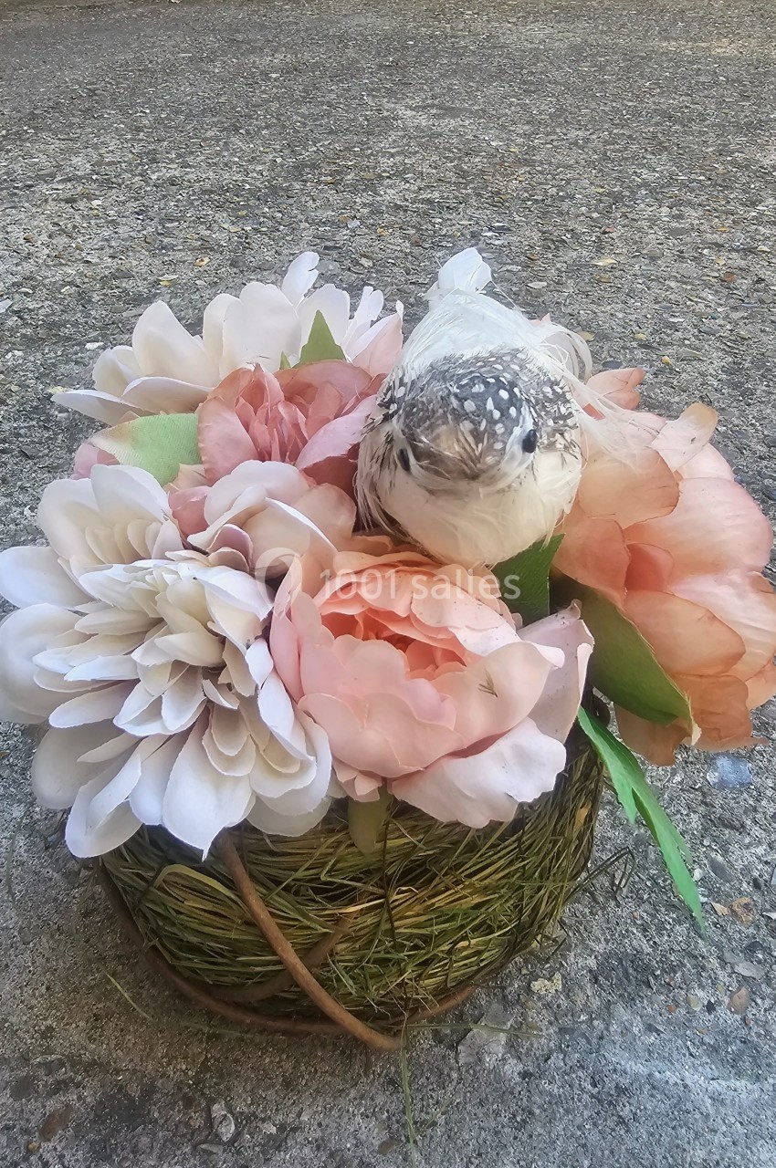 Un petit oiseau perché sur un bouquet de fleurs roses et blanches dans un panier en osier, posé sur un sol en béton.