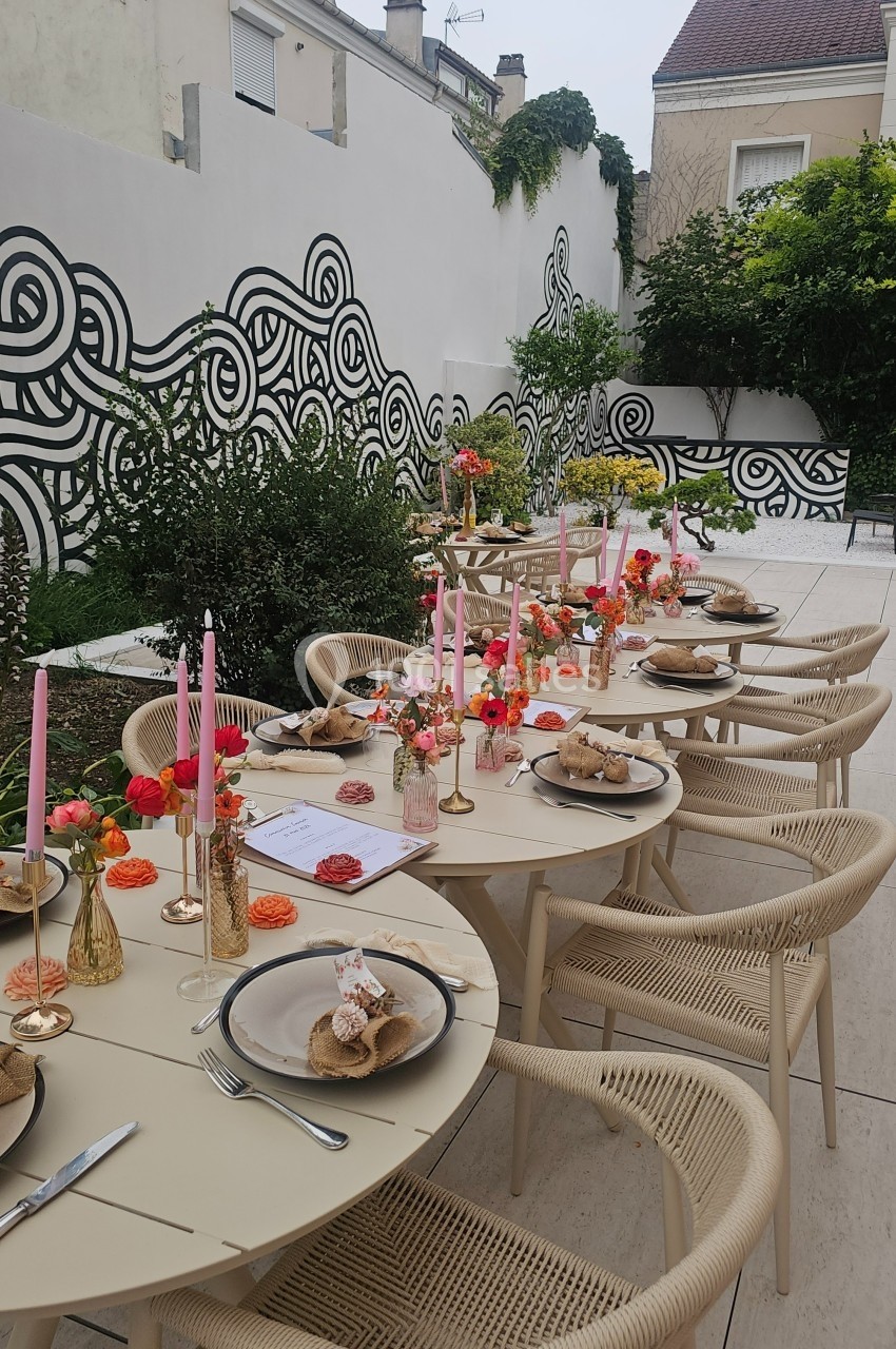Table dressée en extérieur avec des assiettes, bougies roses et décorations florales, entourée de murs peints de motifs…