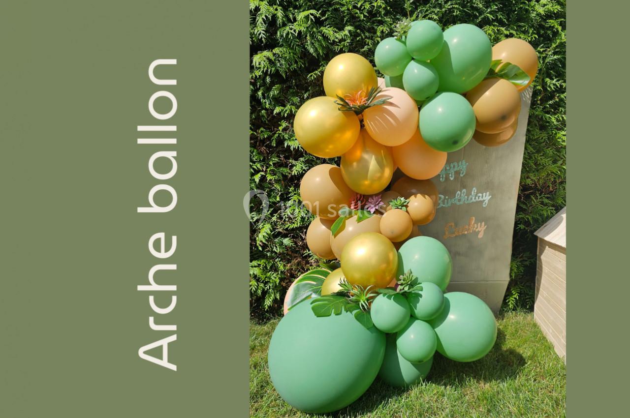 Arche décorative en ballons verts et dorés avec des fleurs, placée devant un panneau en bois dans un jardin.