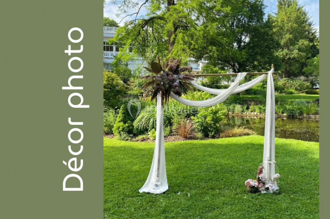 Arche décorative en bois avec voilages blancs et fleurs, installée sur une pelouse devant un jardin verdoyant.