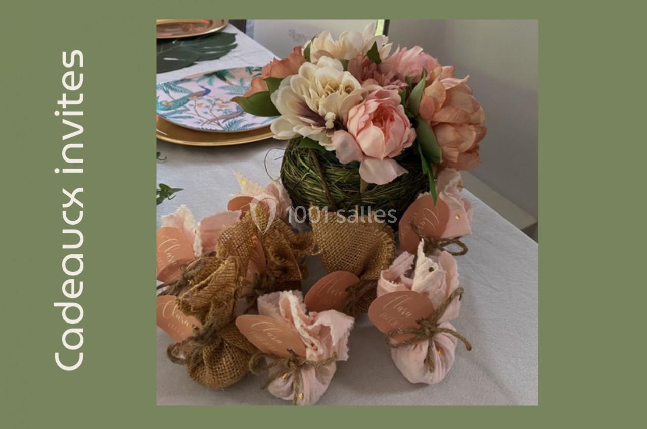 Arrangement floral avec des fleurs roses et blanches, entouré de petits sachets cadeaux en tissu et jute sur une table.