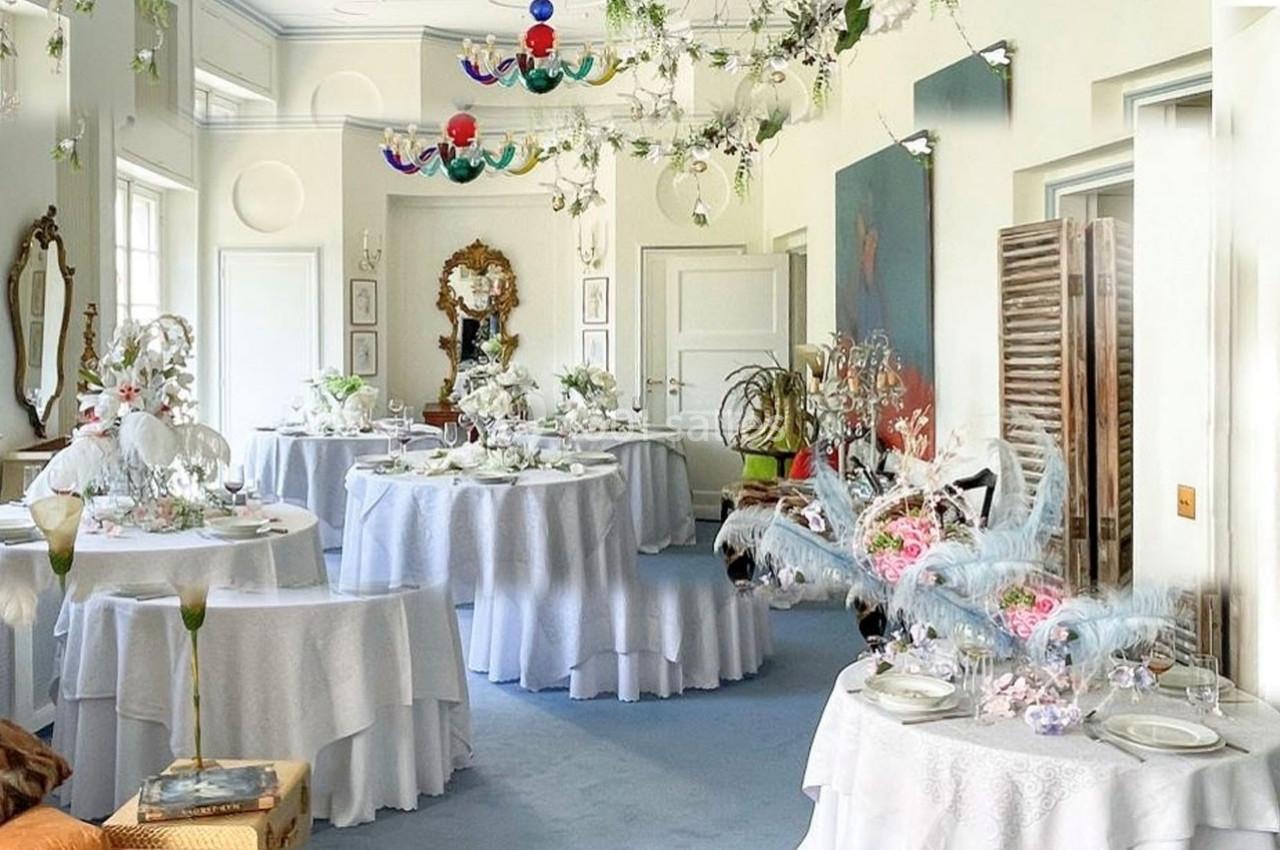 Salle lumineuse décorée pour un événement, avec des tables rondes dressées et des ornements floraux suspendus.