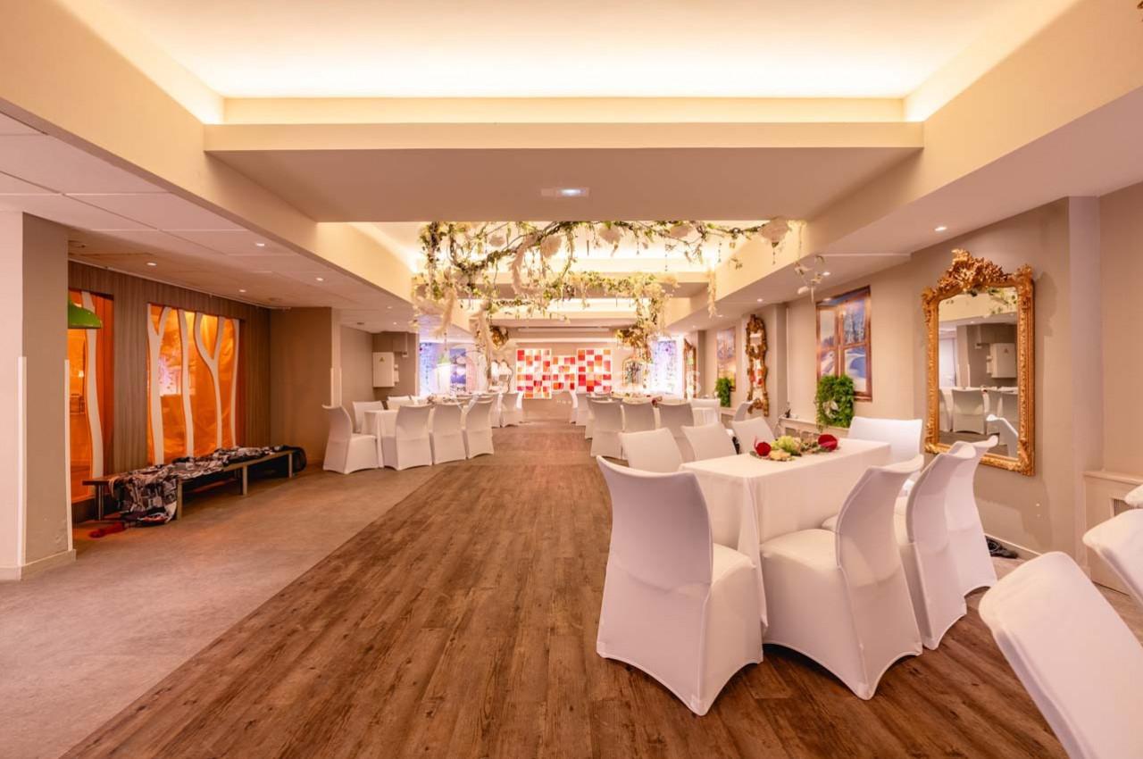 Salle lumineuse avec tables et chaises blanches, décorations florales suspendues et sol en parquet.