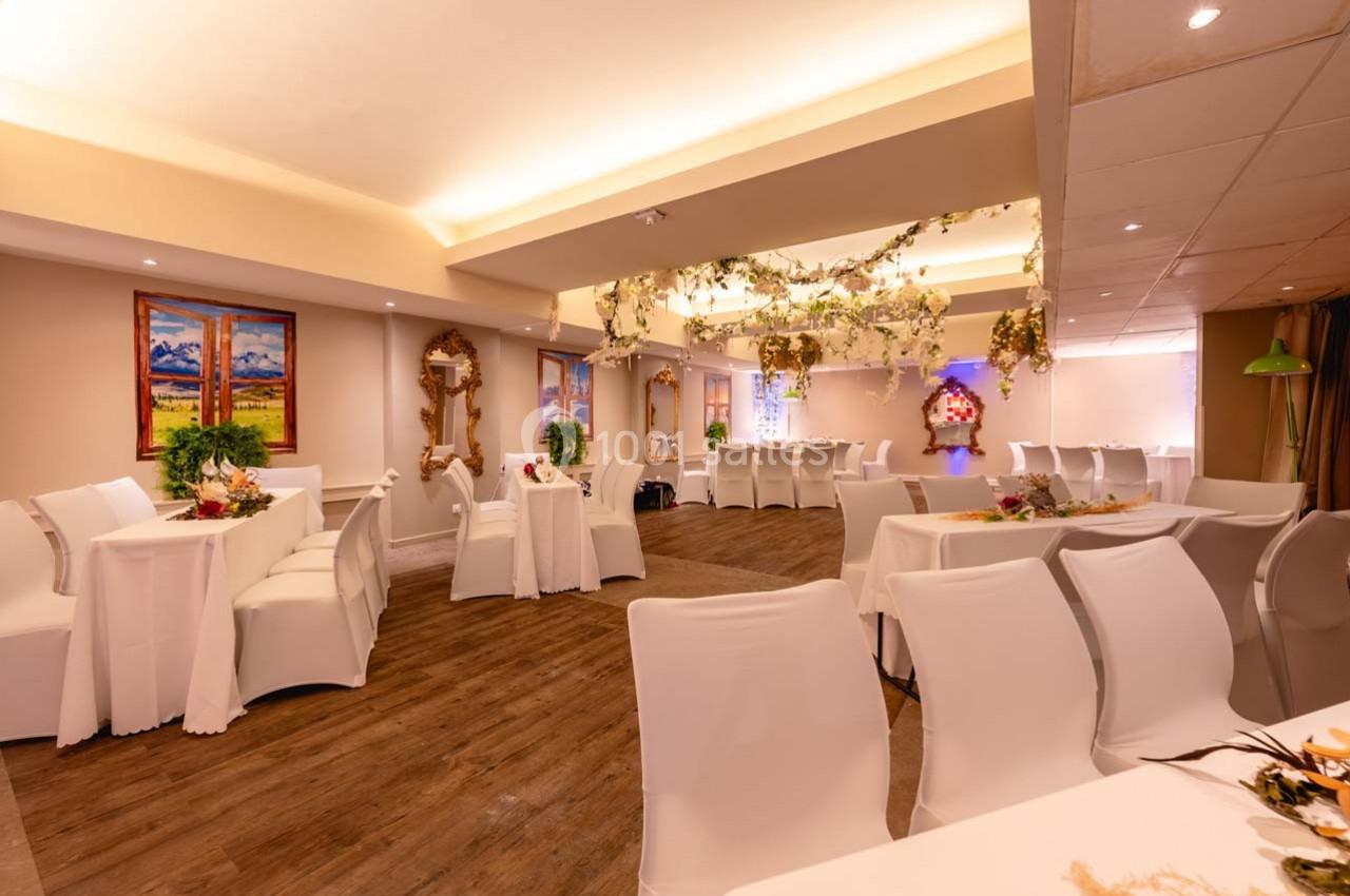 Salle de réception lumineuse avec tables et chaises blanches, décorations florales suspendues et tableaux aux murs.