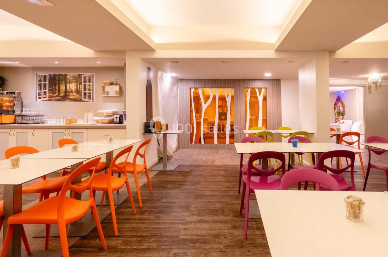 Salle lumineuse avec tables blanches, chaises colorées et grandes baies vitrées décorées de motifs d'arbres.