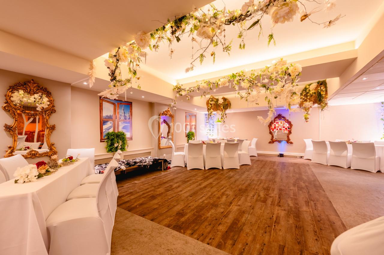 Salle décorée pour un événement, avec chaises blanches, tables dressées, guirlandes florales et miroirs dorés.