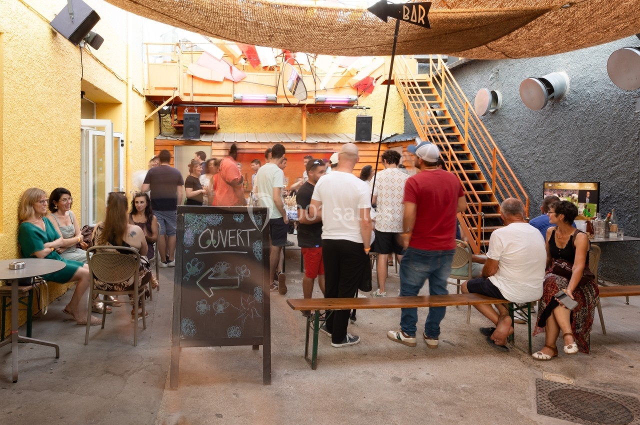Des personnes discutent et se détendent dans une cour extérieure avec des tables, un tableau noir et un escalier en arrière…