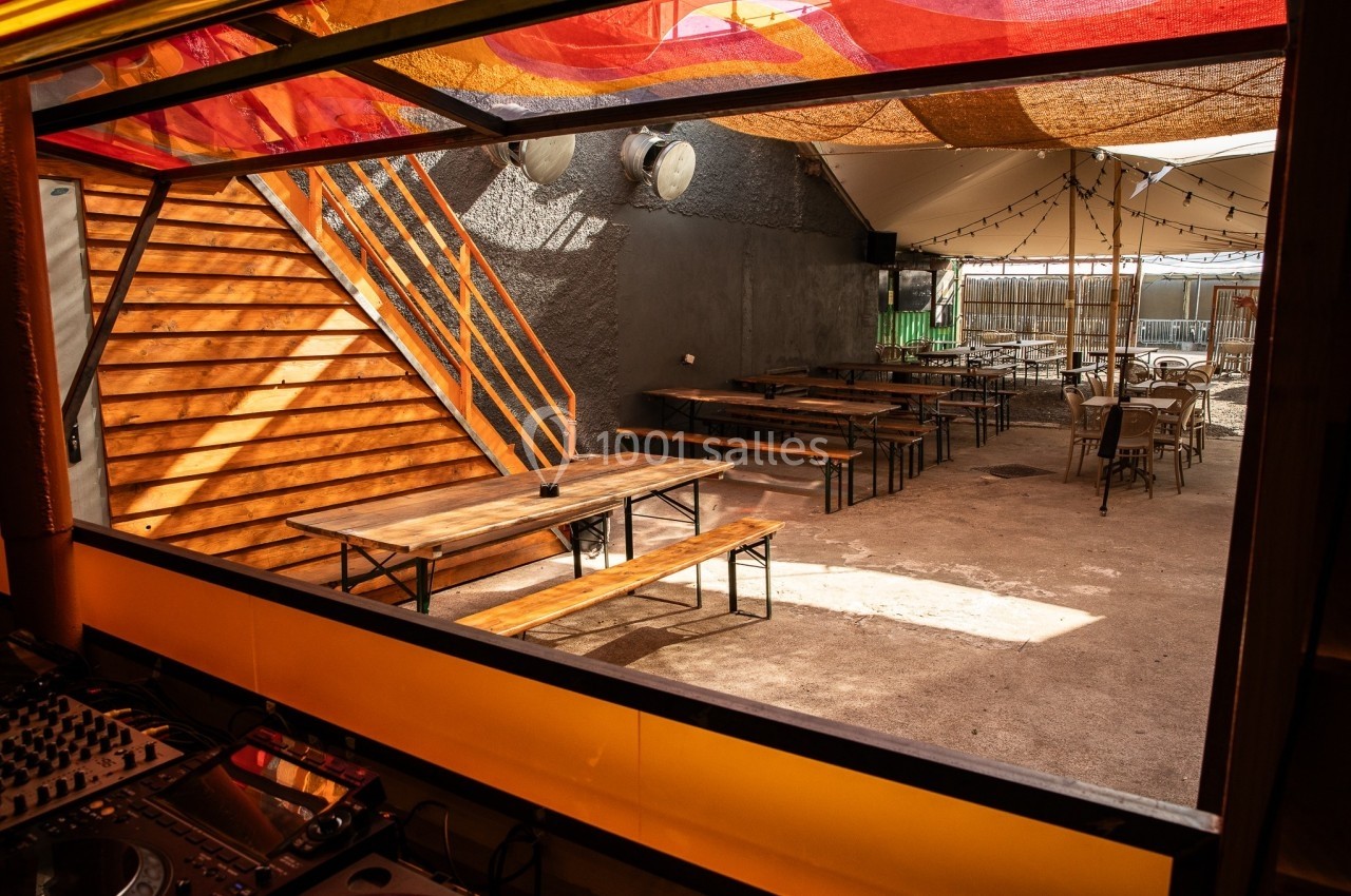 Terrasse extérieure avec tables en bois, bancs, escaliers en bois et toit partiellement couvert de toiles colorées.