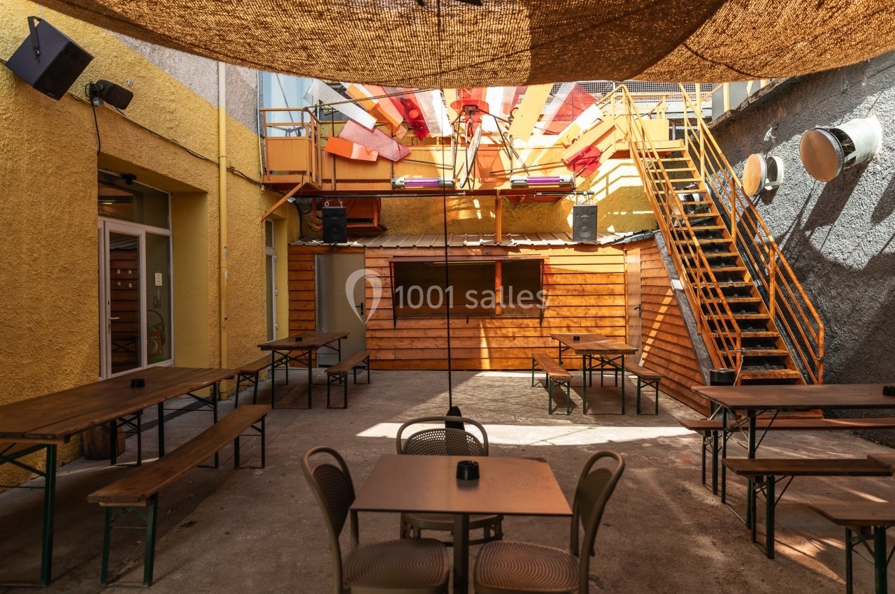 Cour intérieure avec tables en bois, bancs, escalier métallique et décorations colorées sous une toile d'ombrage.