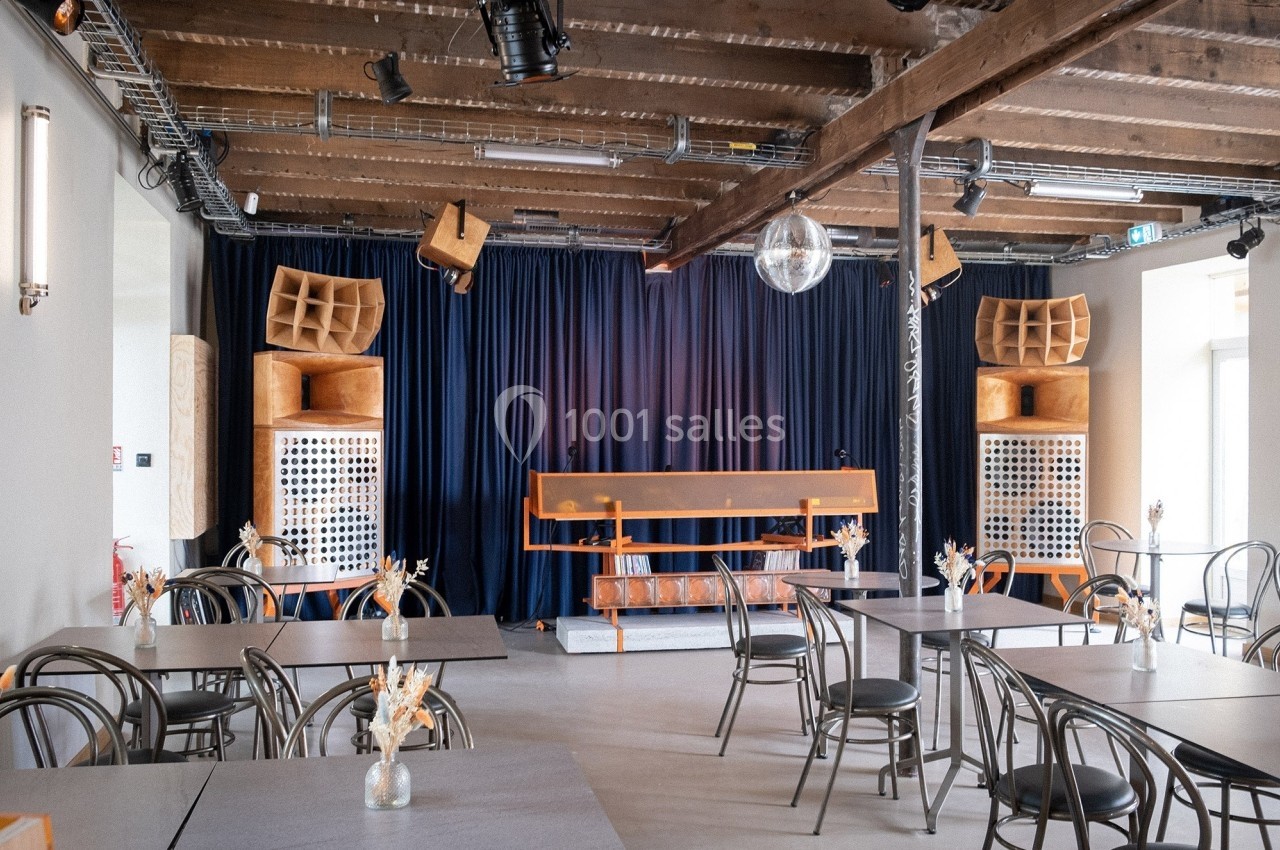 Salle avec tables, chaises, scène équipée de matériel audio, rideaux bleus et plafond en poutres apparentes.