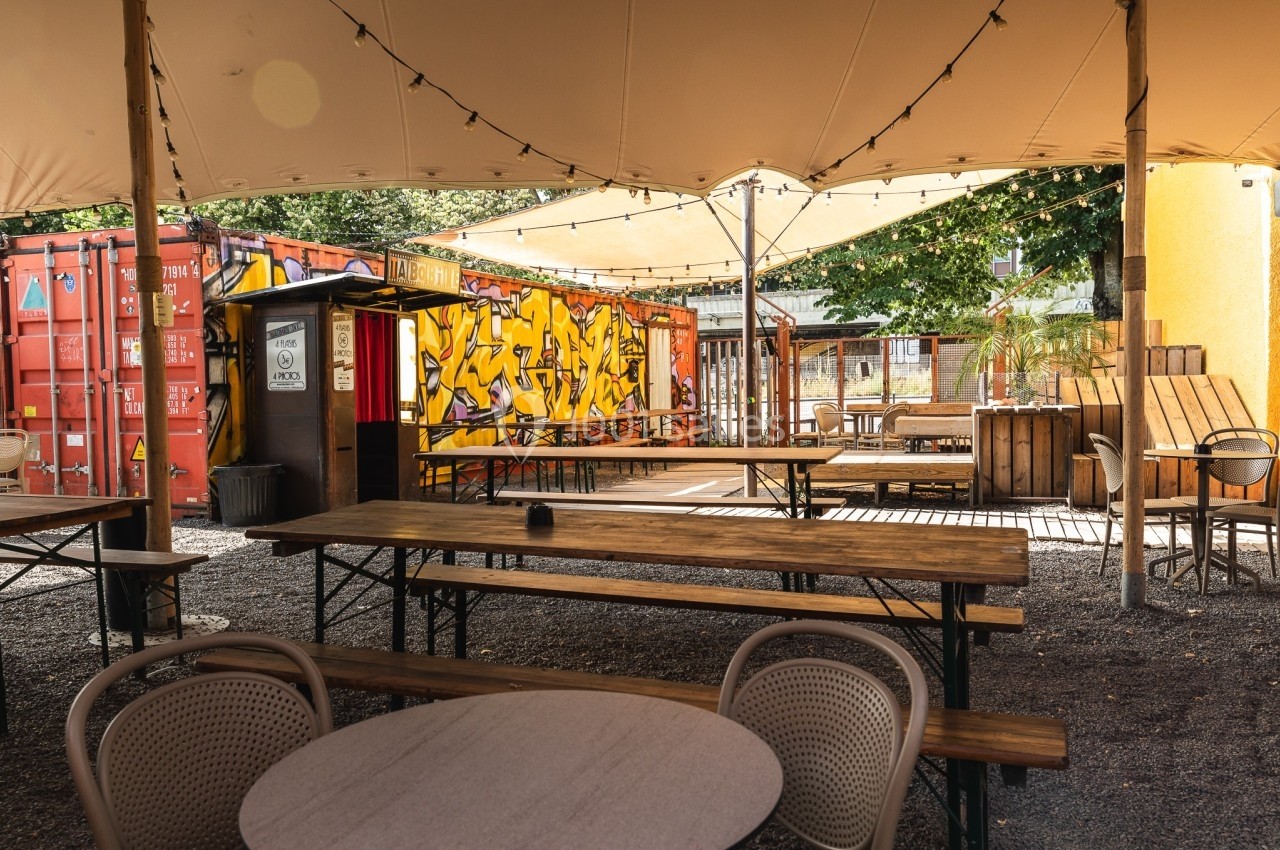 Terrasse extérieure avec tables en bois, chaises en rotin, conteneurs colorés et graffitis en arrière-plan.