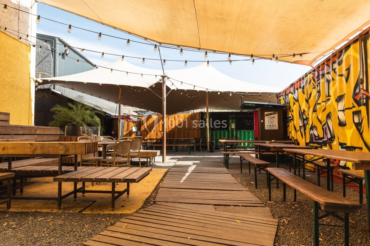 Terrasse extérieure avec tables en bois, guirlandes lumineuses suspendues et mur décoré de graffitis colorés.