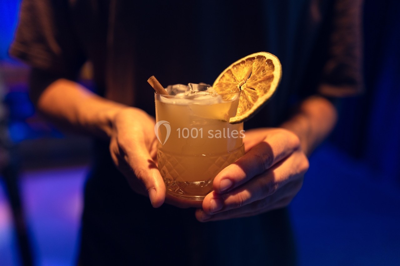 Un verre de cocktail avec une tranche d'orange séchée et une paille, tenu par une personne dans un éclairage tamisé.