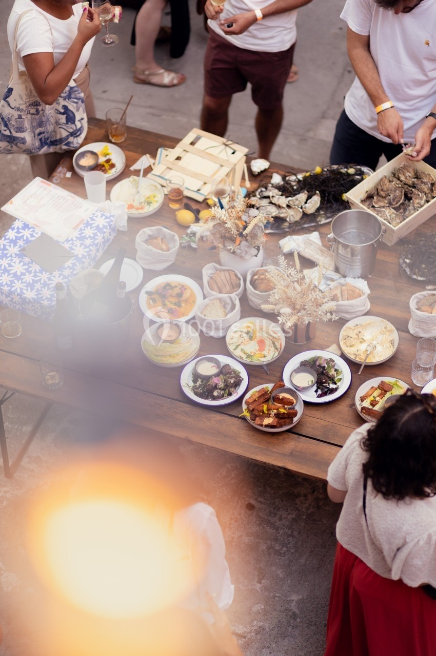 Table en bois garnie de plats variés, fruits de mer, pains et boissons, entourée de convives debout.