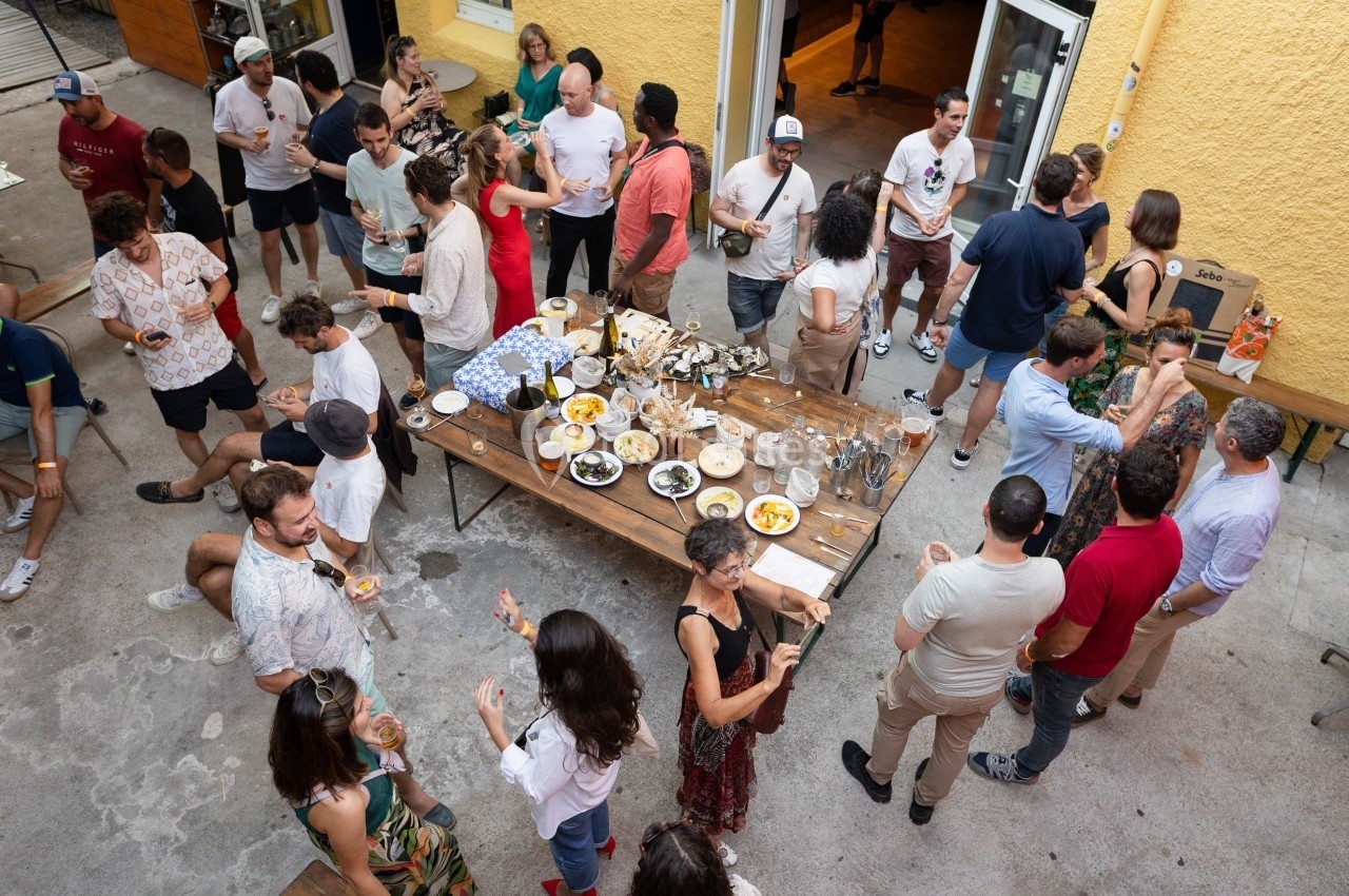 Groupe de personnes discutant et partageant un repas autour d'une grande table dans une cour extérieure.