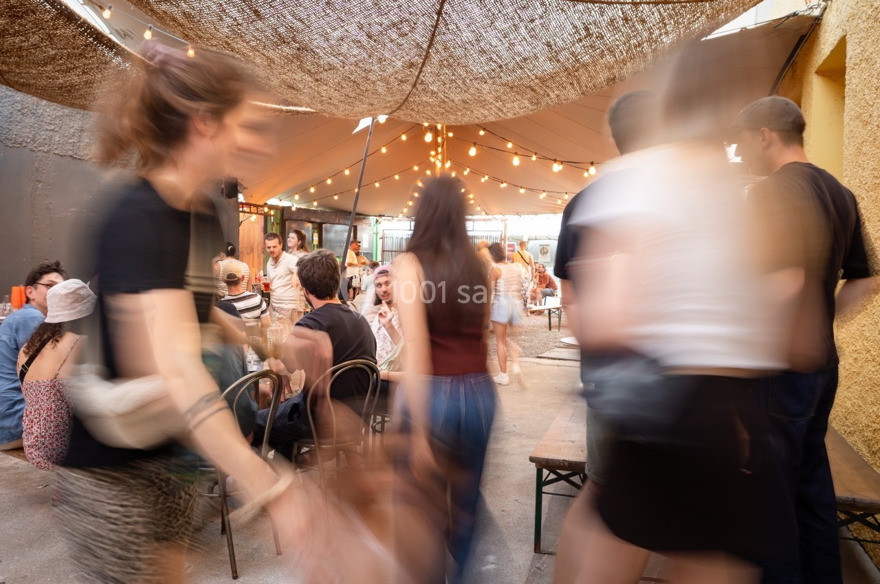 Personnes se déplaçant dans un espace extérieur animé, avec des tables, des chaises et des guirlandes lumineuses suspendues.