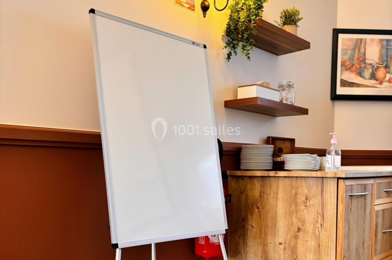 Tableau blanc sur trépied dans une pièce avec étagères, vaisselle, plantes et un meuble en bois.