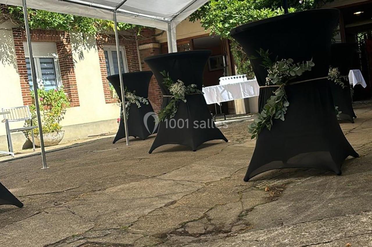 Tables hautes noires décorées de feuillages, disposées en extérieur sous une tente blanche, près d'un bâtiment en briques.