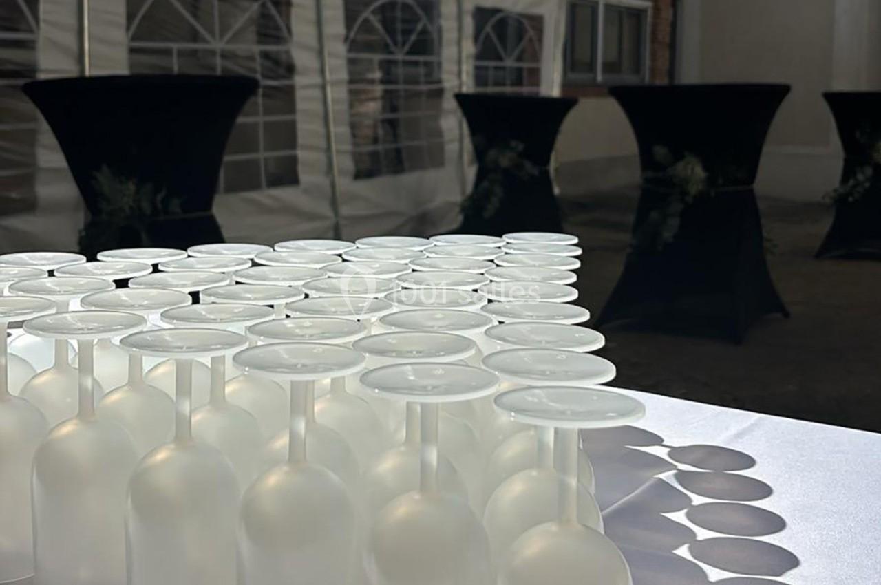 Rangée de verres à pied retournés sur une table blanche, avec des tables hautes en arrière-plan sous une tente.