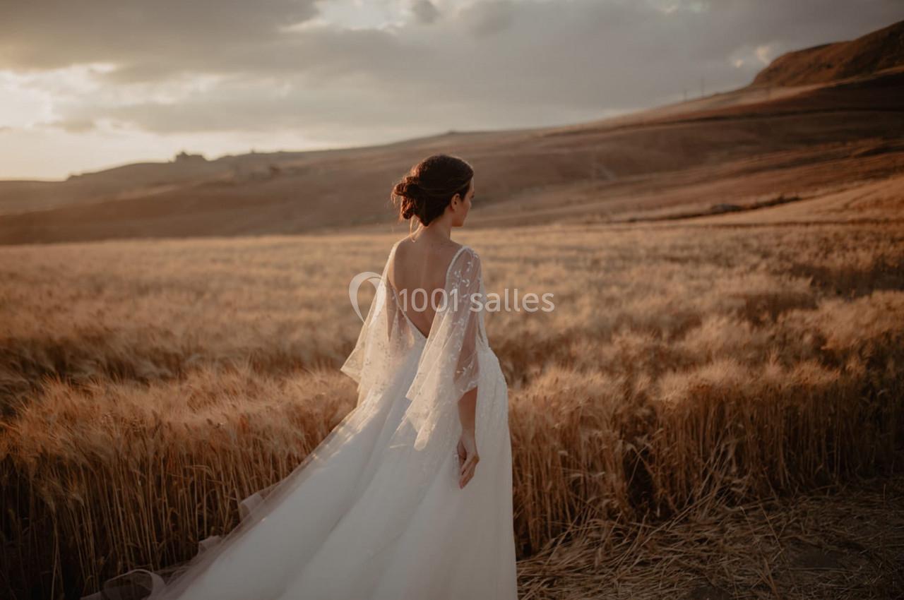 Une femme en robe de mariée marche dans un champ de blé au coucher du soleil, sous un ciel partiellement nuageux.
