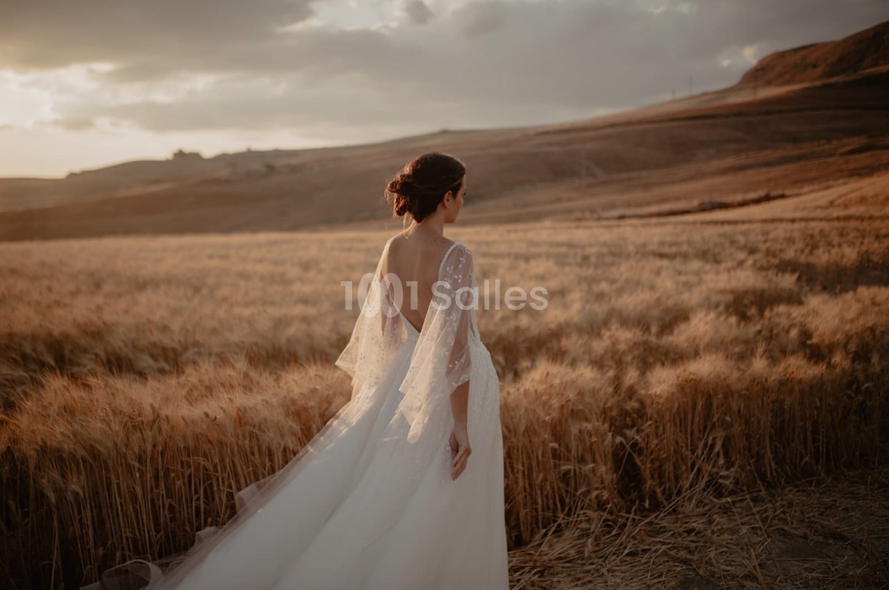 Une femme en robe de mariée marche dans un champ de blé au coucher du soleil, sous un ciel partiellement nuageux.