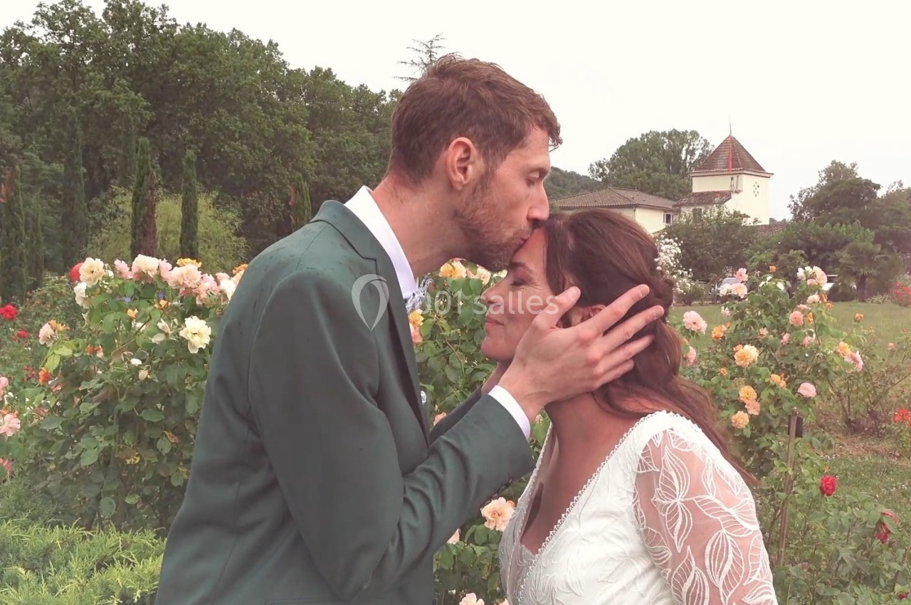 Un homme en costume vert embrasse tendrement le front d'une femme en robe blanche dans un jardin fleuri.