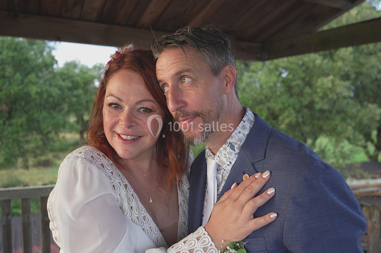Un couple souriant en tenue de mariage pose sous un abri en bois, avec un paysage verdoyant en arrière-plan.