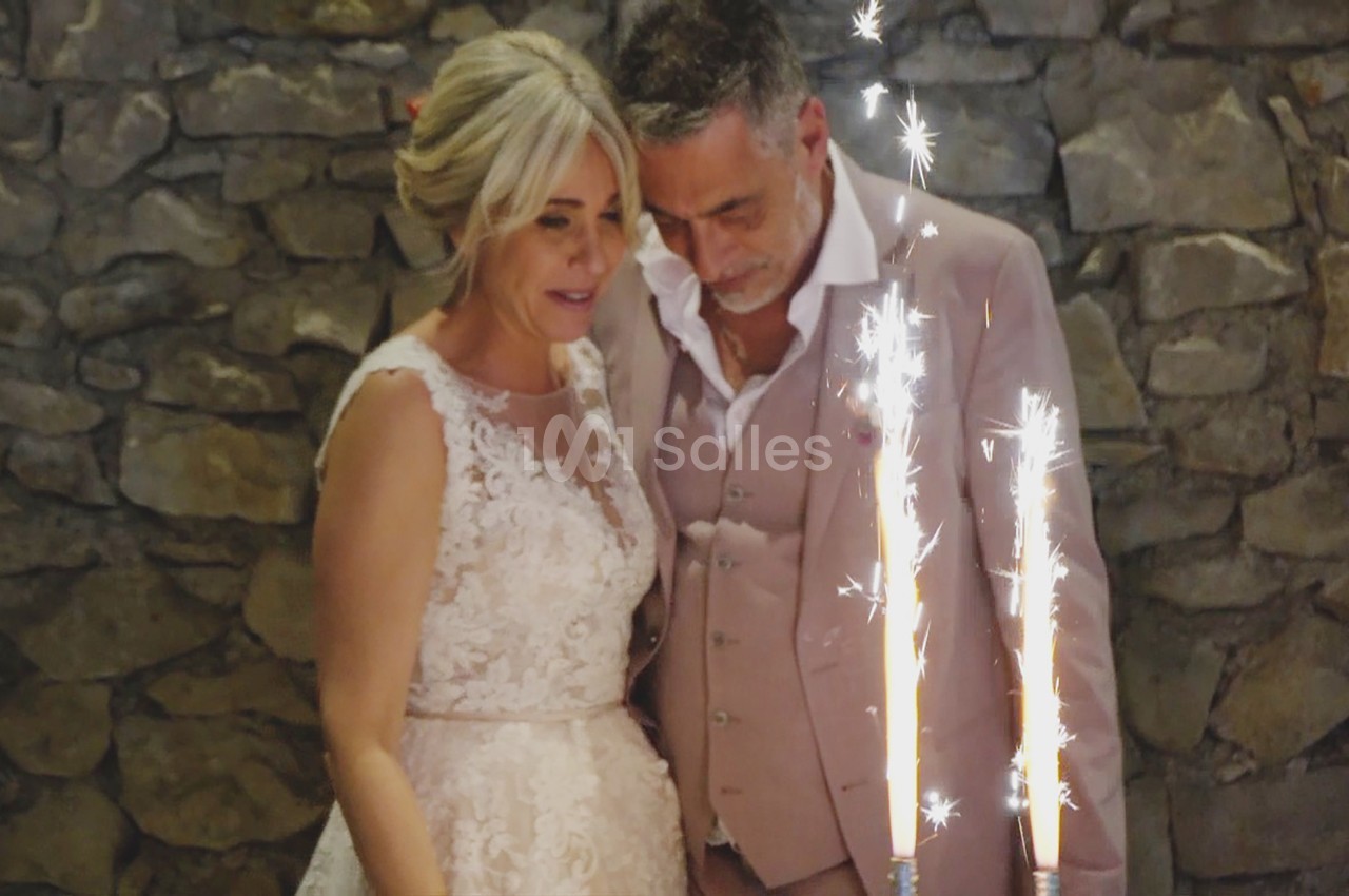 Un couple en tenue de mariage regarde des fontaines lumineuses devant un mur en pierre.