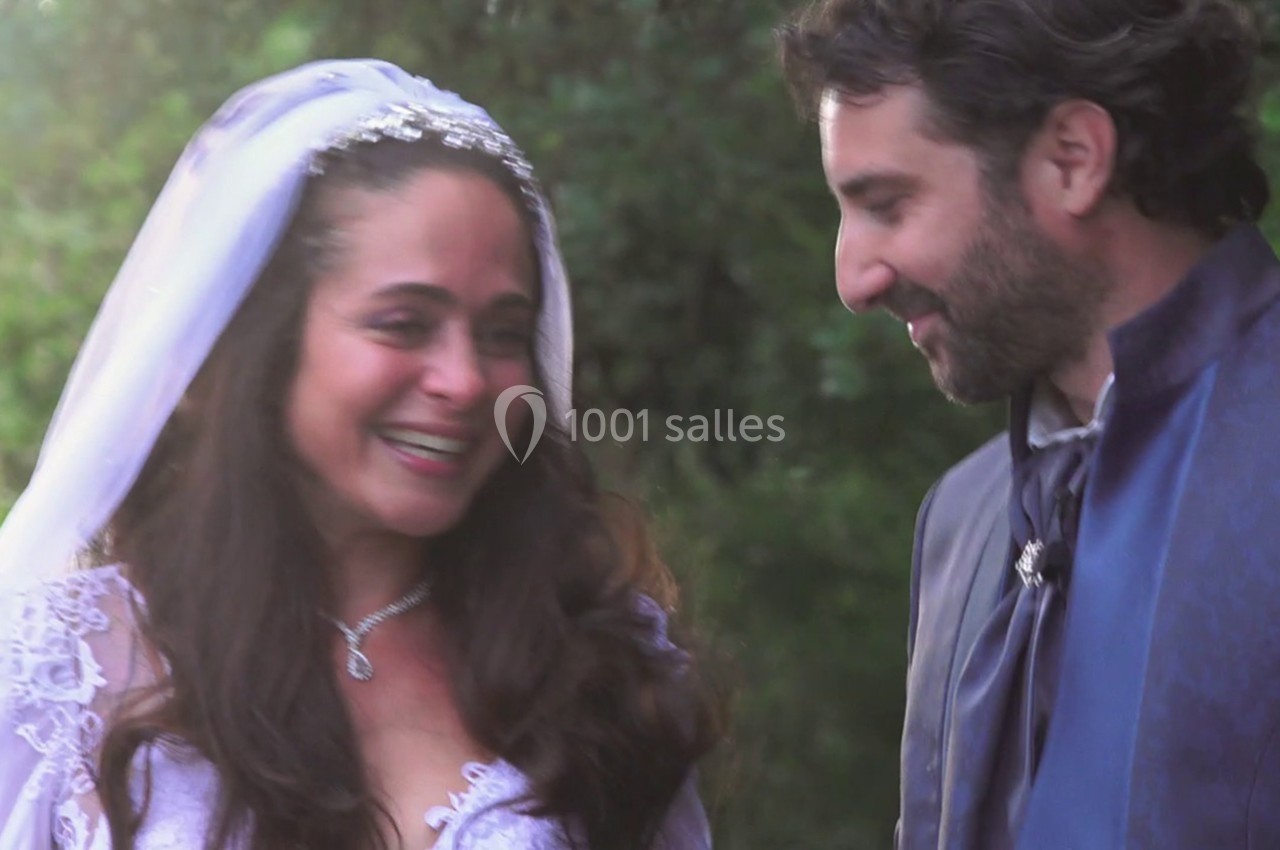 Une mariée souriante en robe blanche et voile discute avec un homme en costume dans un cadre extérieur verdoyant.