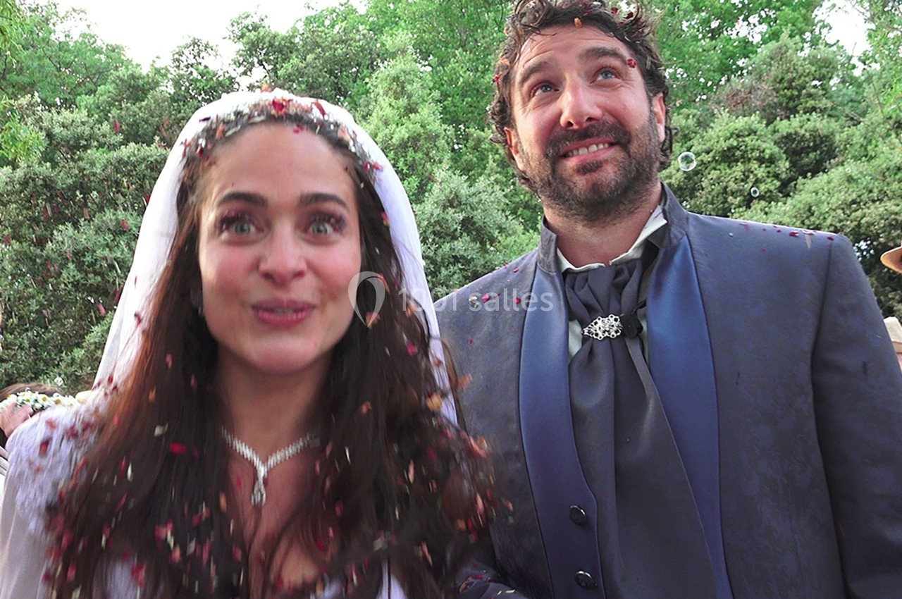 Un couple souriant en tenue de mariage, couvert de pétales, devant un arrière-plan verdoyant.