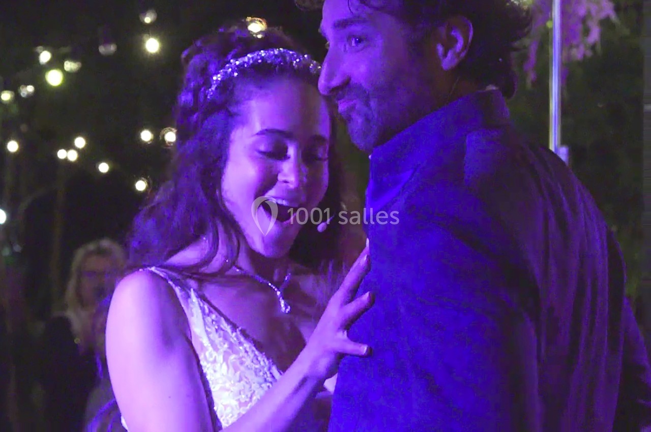 Une femme en robe blanche sourit en touchant un homme en costume sous des lumières tamisées en extérieur.