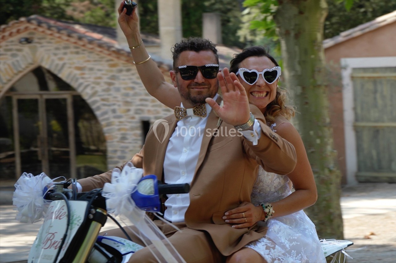 Un couple en tenue de mariage sourit et salue en roulant sur une moto décorée, dans un cadre extérieur ensoleillé.