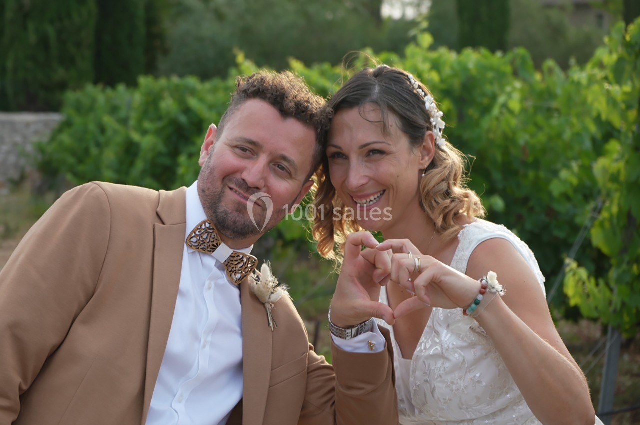 Un couple souriant en tenue de mariage formant un cœur avec leurs mains devant un décor naturel verdoyant.
