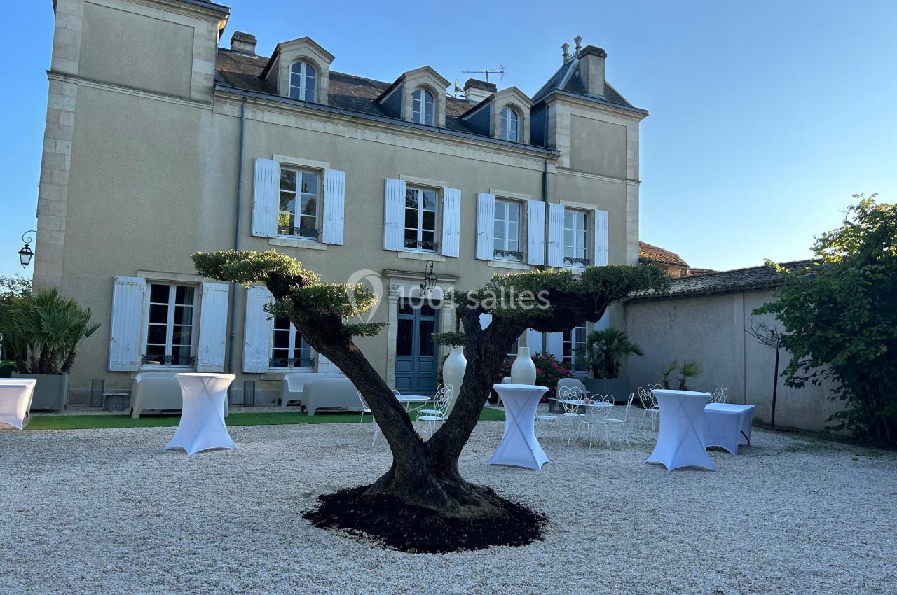 Façade d'une maison en pierre avec un arbre taillé au premier plan et des tables dressées sur une cour gravillonnée.