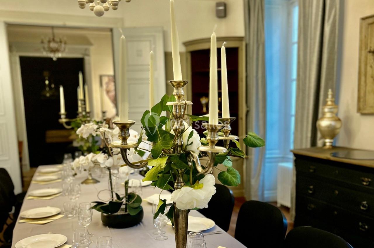 Table élégamment dressée avec chandeliers dorés, fleurs blanches et vaisselle, dans une salle lumineuse et raffinée.