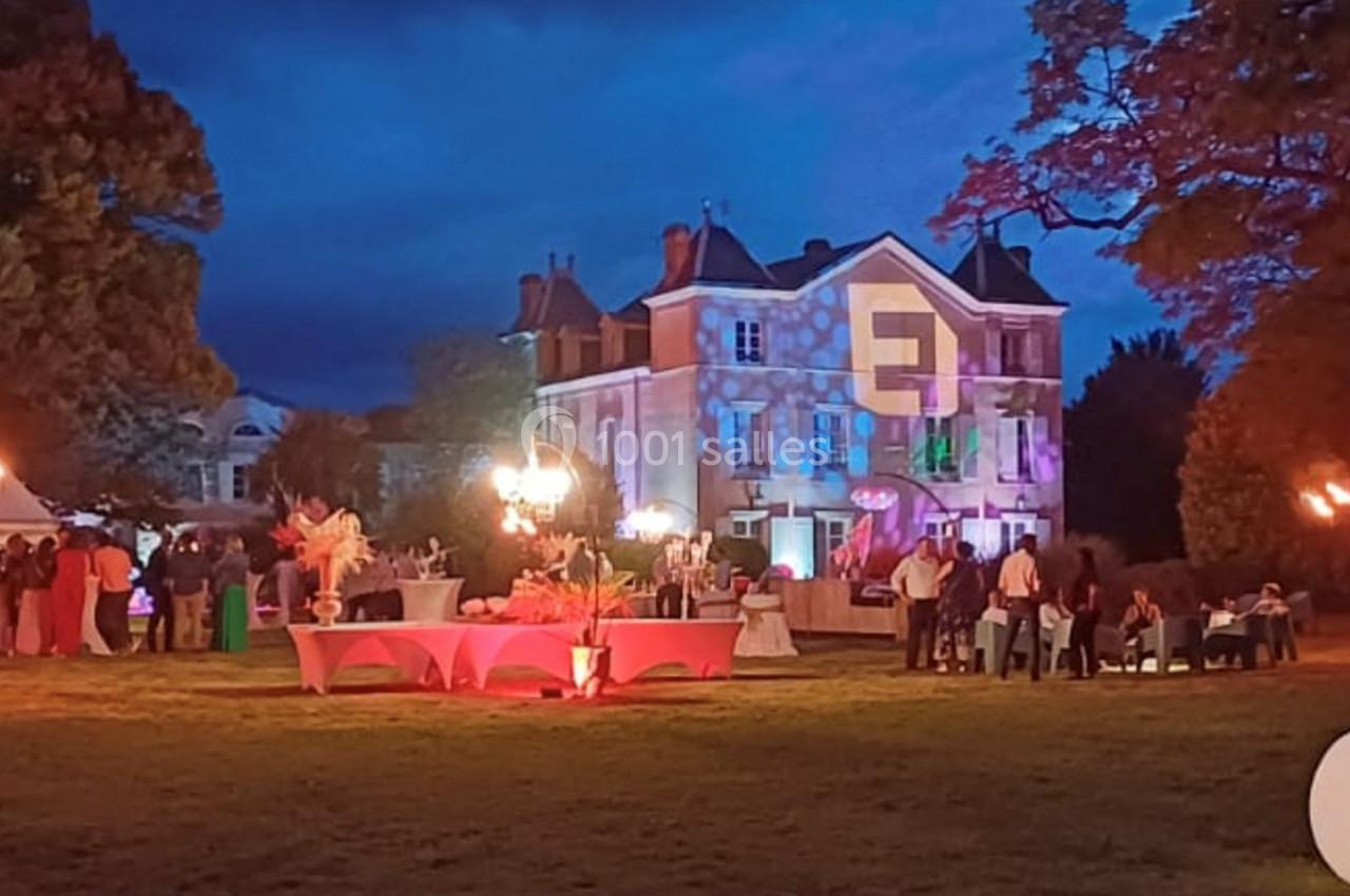Manoir illuminé de nuit avec des décorations lumineuses, des tables dressées et des invités dans un jardin.