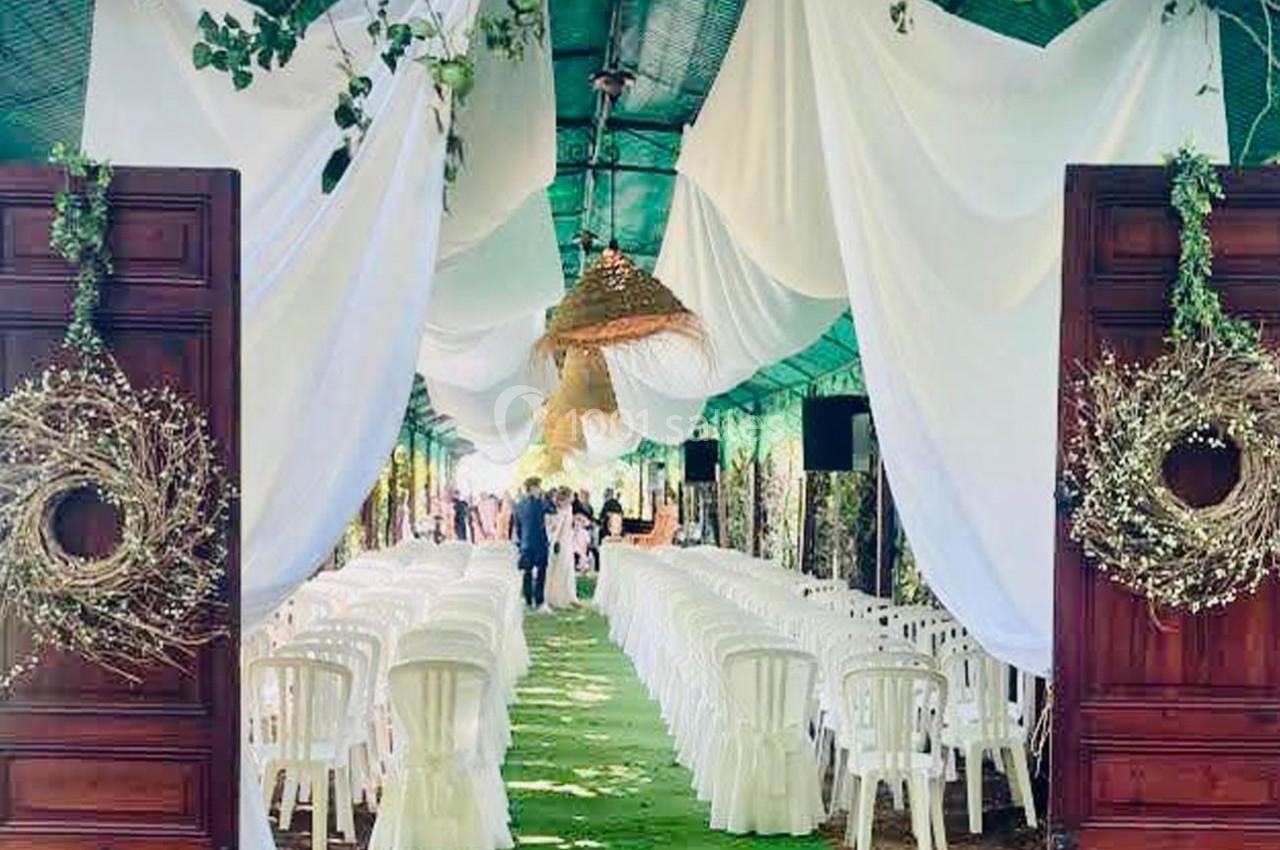 Allée décorée pour une cérémonie, avec chaises blanches alignées, drapés suspendus et végétation environnante.