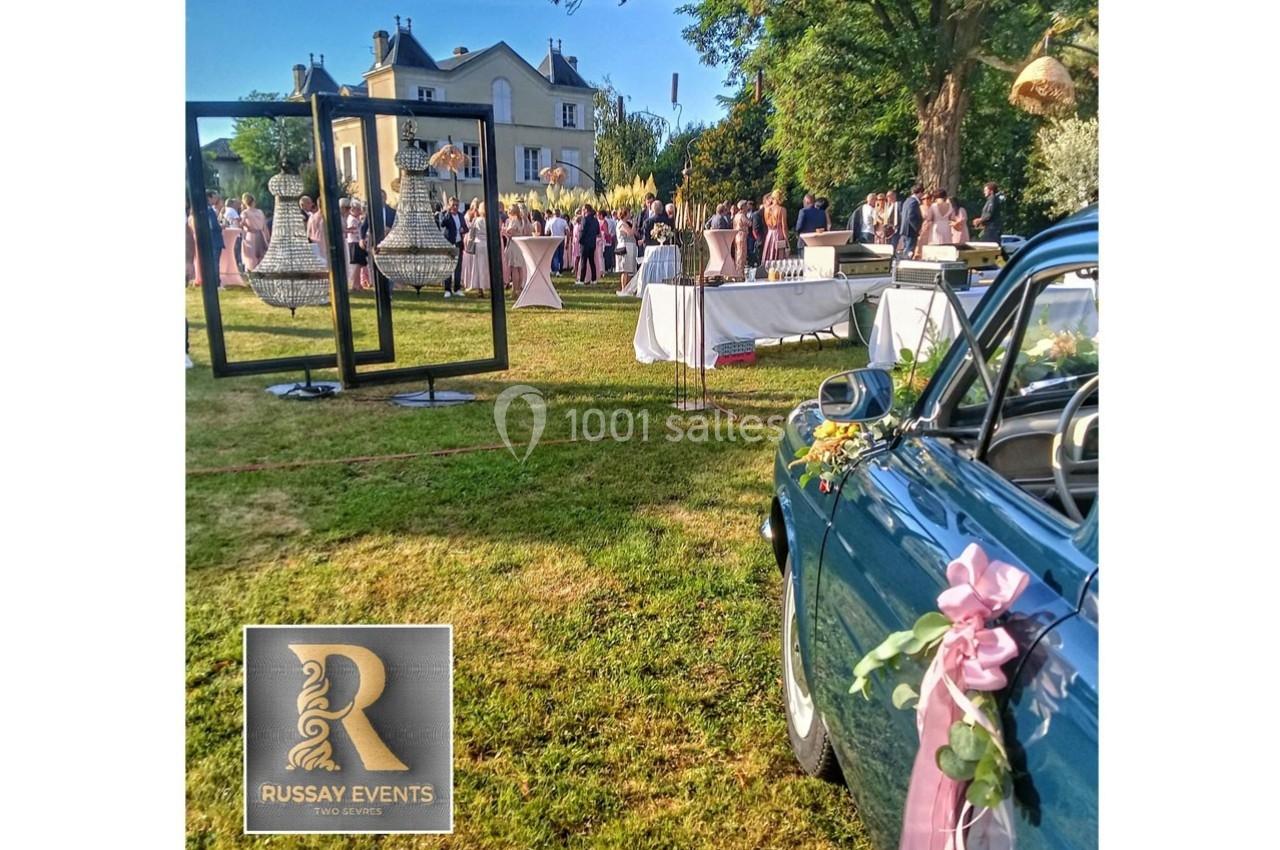 Vue d'un événement en plein air avec invités, décorations élégantes, voiture ancienne et un bâtiment en arrière-plan.