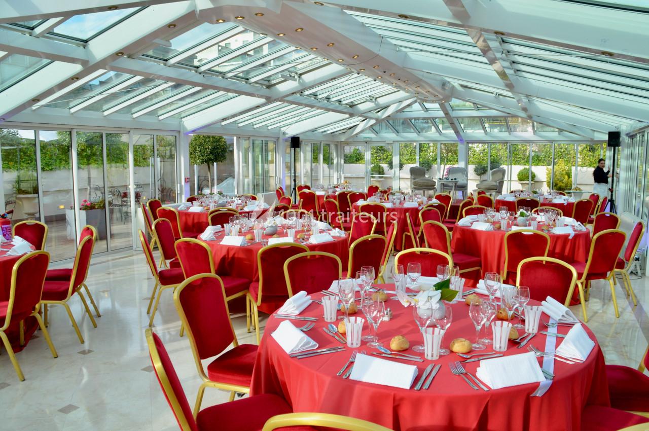 Salle lumineuse avec verrière, tables rondes dressées avec nappes rouges, chaises assorties et couverts élégants.