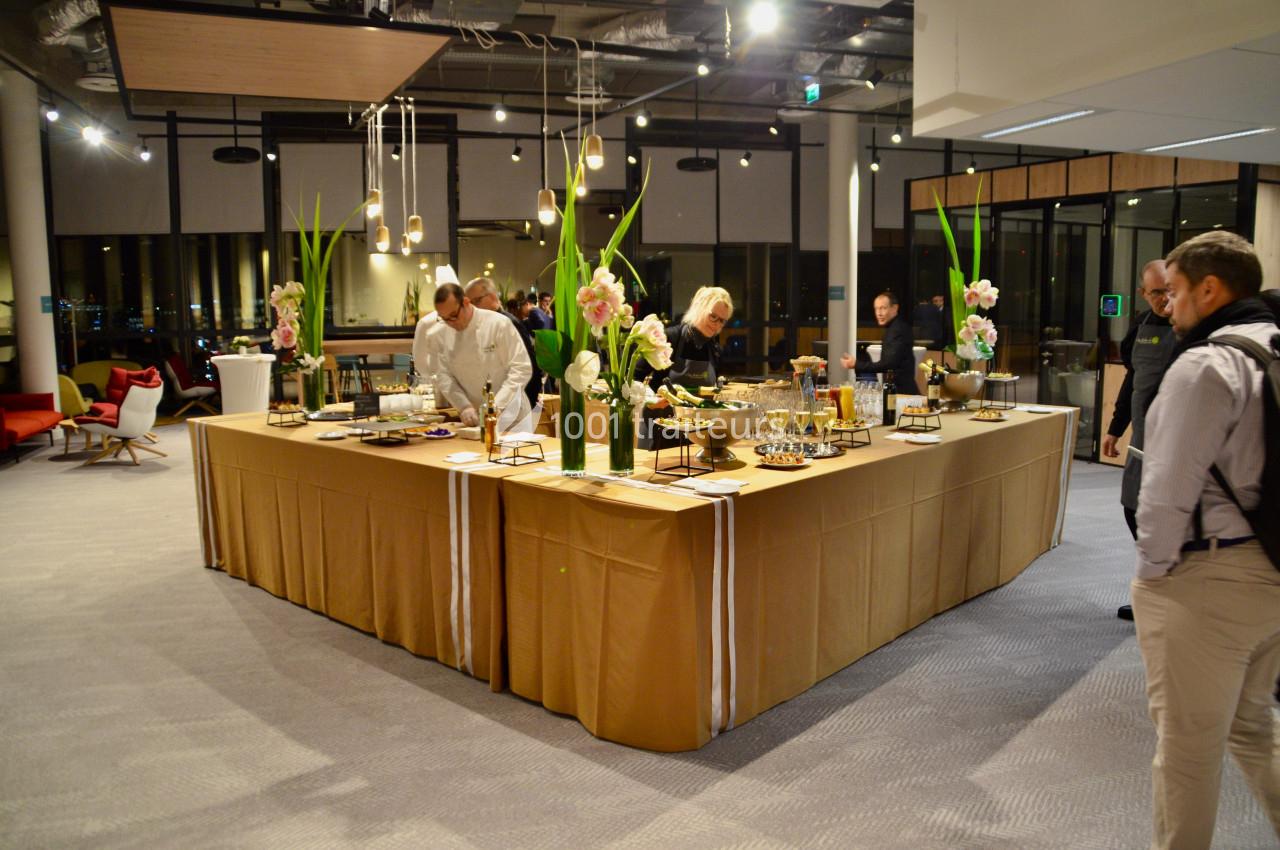Buffet élégant avec nappes dorées, décor floral et invités se servant dans une salle moderne et lumineuse.