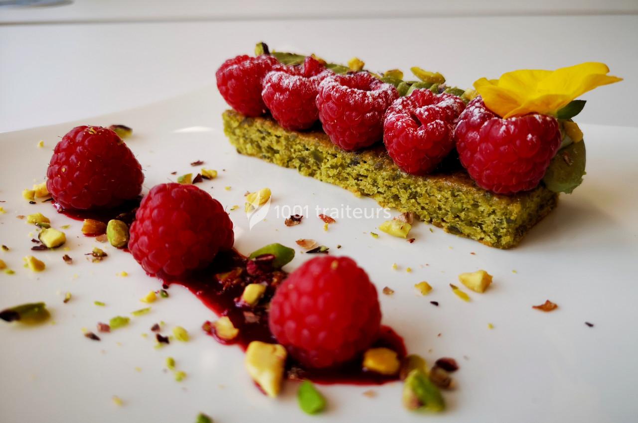 Dessert composé de framboises fraîches sur un biscuit pistache, décoré de pistaches concassées et d'une fleur comestible.