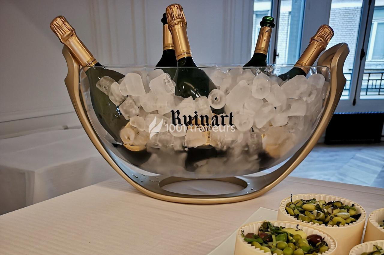Seau en verre rempli de glace contenant des bouteilles de champagne Ruinart, posé sur une table avec des amuse-bouches.