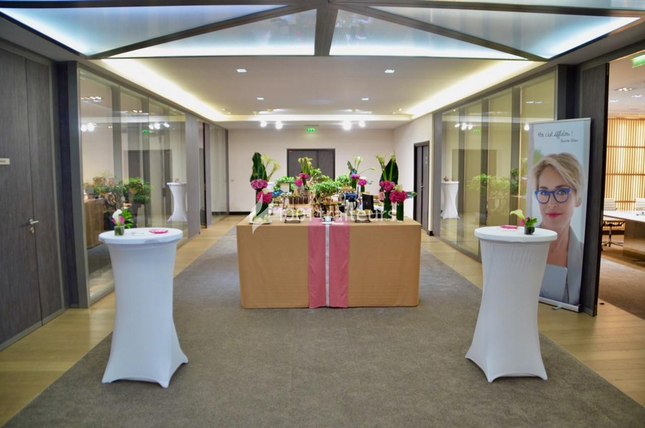 Table décorée avec des fleurs et des boissons dans un hall lumineux, entourée de murs vitrés et de plantes.