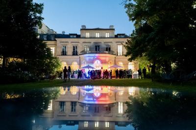 Façade illuminée d'un bâtiment élégant au crépuscule, avec un jardin et des invités rassemblés devant.