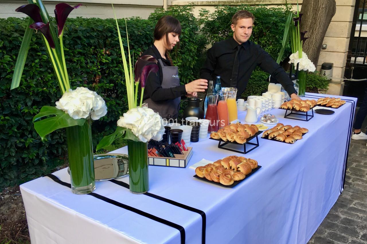 Table de buffet en extérieur avec viennoiseries, jus de fruits, café et fleurs, servie par deux personnes.