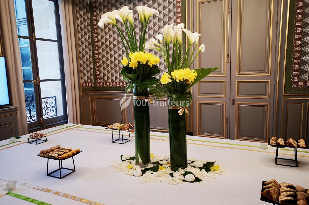 Arrangement floral avec des arums blancs et des fleurs jaunes dans des vases verts, entouré de desserts sur une table…